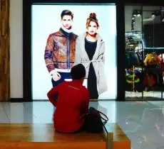 A person sits on a bench looking at a large, bright digital advertisement featuring two fashion models