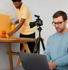 A person sits at a laptop in the foreground, while another prepares orange juice with a camera set up in the background.