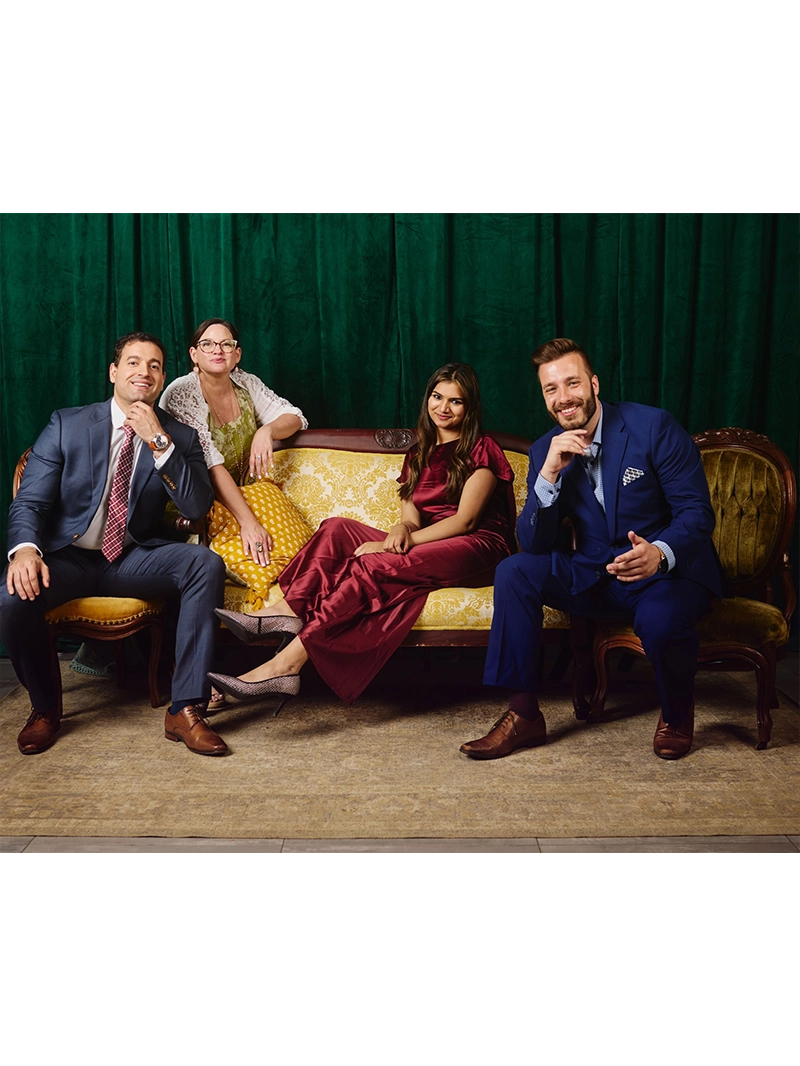 Elegantly dressed individuals sit on a vintage couch, posing against a rich green backdrop with a patterned rug beneath.