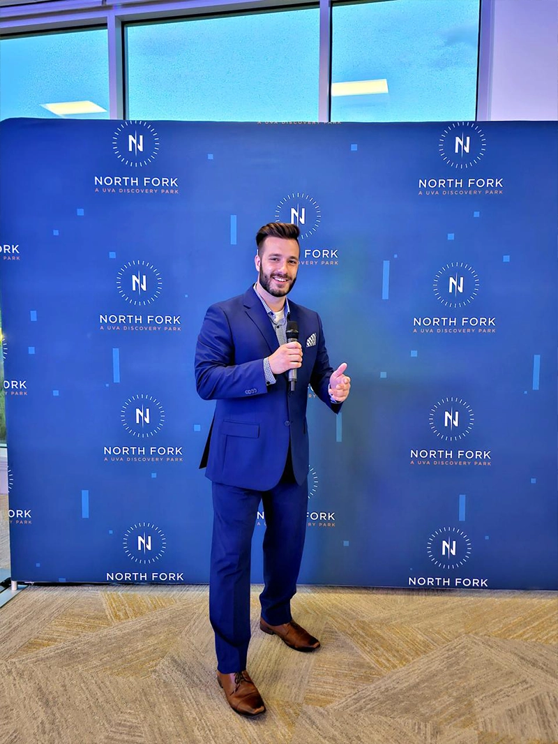 A man in a blue suit stands with a microphone in front of a "North Fork" branded backdrop at a professional event.