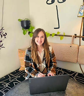 A person sitting at a table with a laptop, wearing a plaid jacket, surrounded by plants and modern decor in a cozy space.
